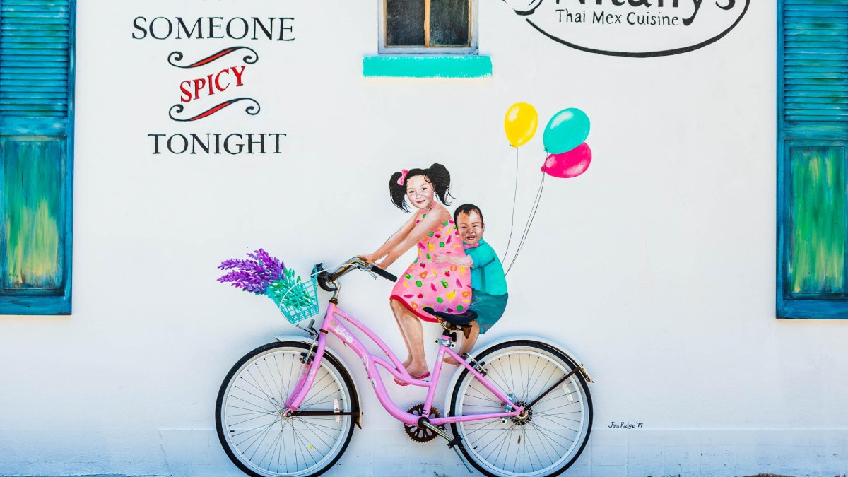 Colorful painting on a white wall of two kids riding a pink bike with balloons on the back