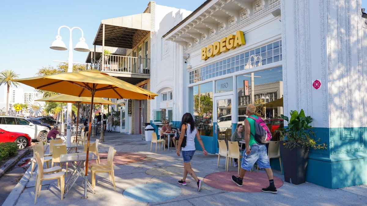 Two people in casual summer clothes walk past bodega in the EDGE District of St. Pete. Tables with yellow umbrellas are on the sidewalk