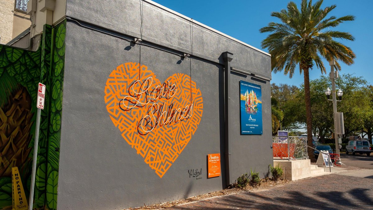 a mural of an orange heart that says Love Shines on a dark gray building with a blue sign and a palm tree to the right