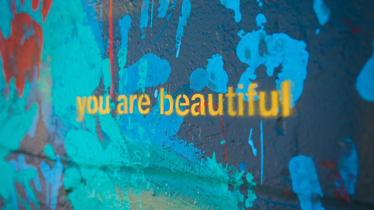 a portion of a large mural displays the words "you are beautiful' in yellow lowercase letters against a background of painted blue, green and red handprints on a dark background