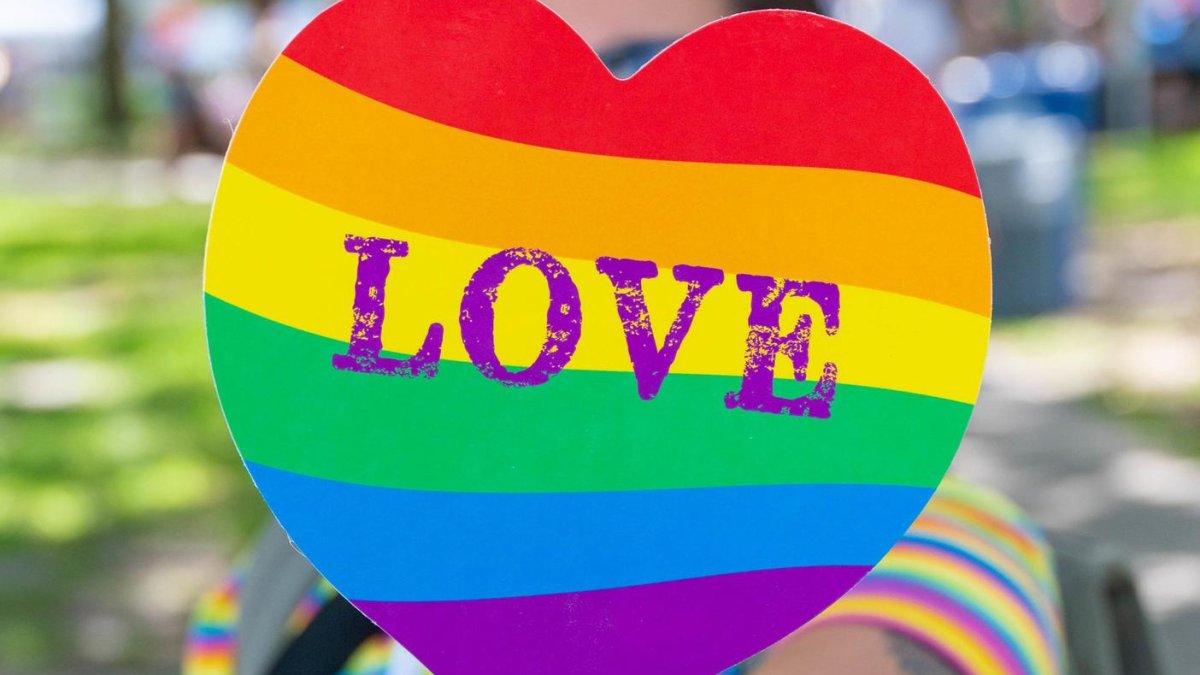A woman in a striped rainbow shirt holds up a heart-shaped rainbow colored fan