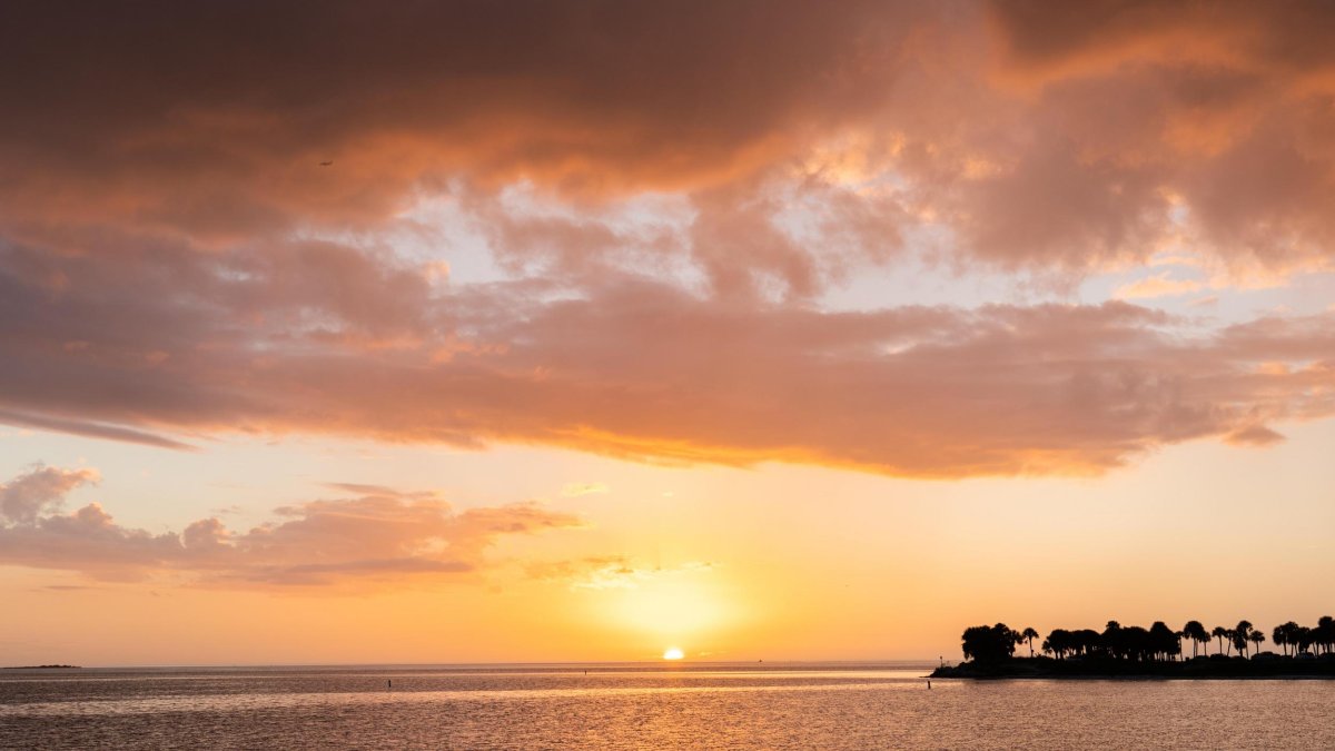 um céu ao pôr do sol com cores amarelas e rosa e uma grande formação de nuvens, a água está em primeiro plano com a terra pode ser vista à direita