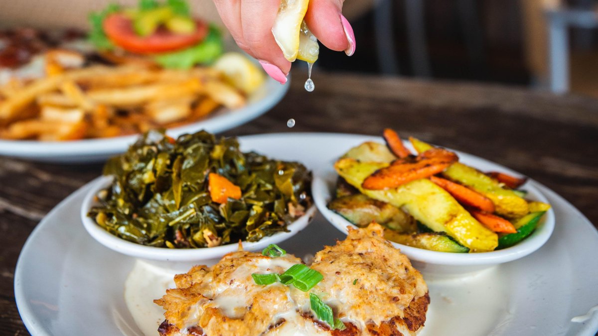 a woman squeezing a lemon on top of a plate of grilled fish, sauteed greens and mixed veg at Hog Island Fish Camp in Dunedin