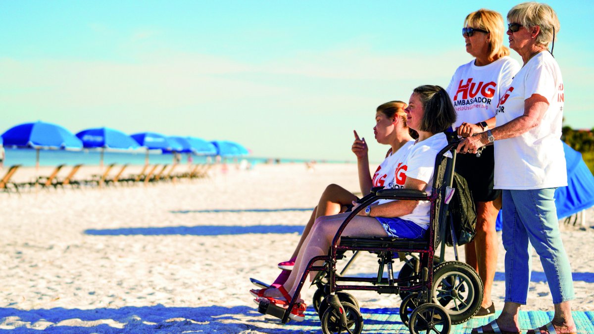 two women in white t-shirts and jeans accommodate two young girls in wheelchairs on a beach access mat in Indian Rock Beach, blue umbrellas are in front of them on the sand