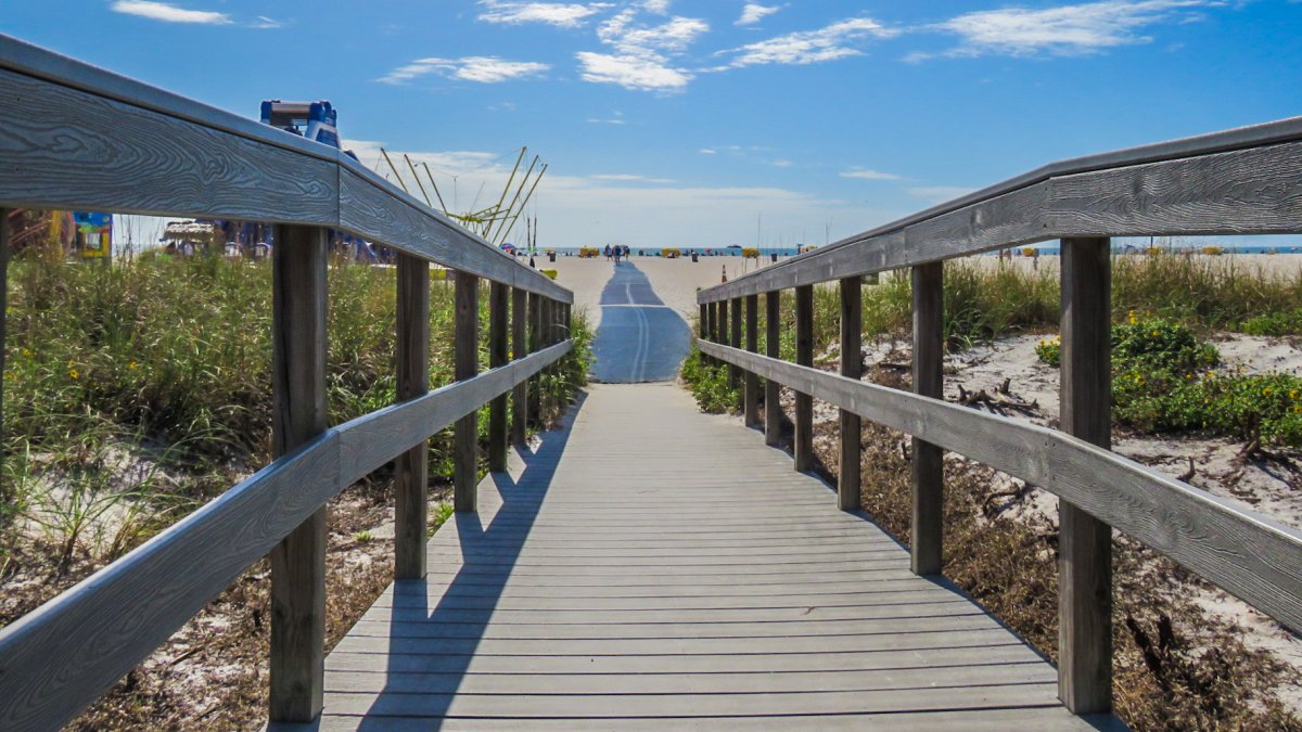 boardwalk to beach lined with dunes, with a large inflatable water slide on the left, the sky is blue with puffy white clouds