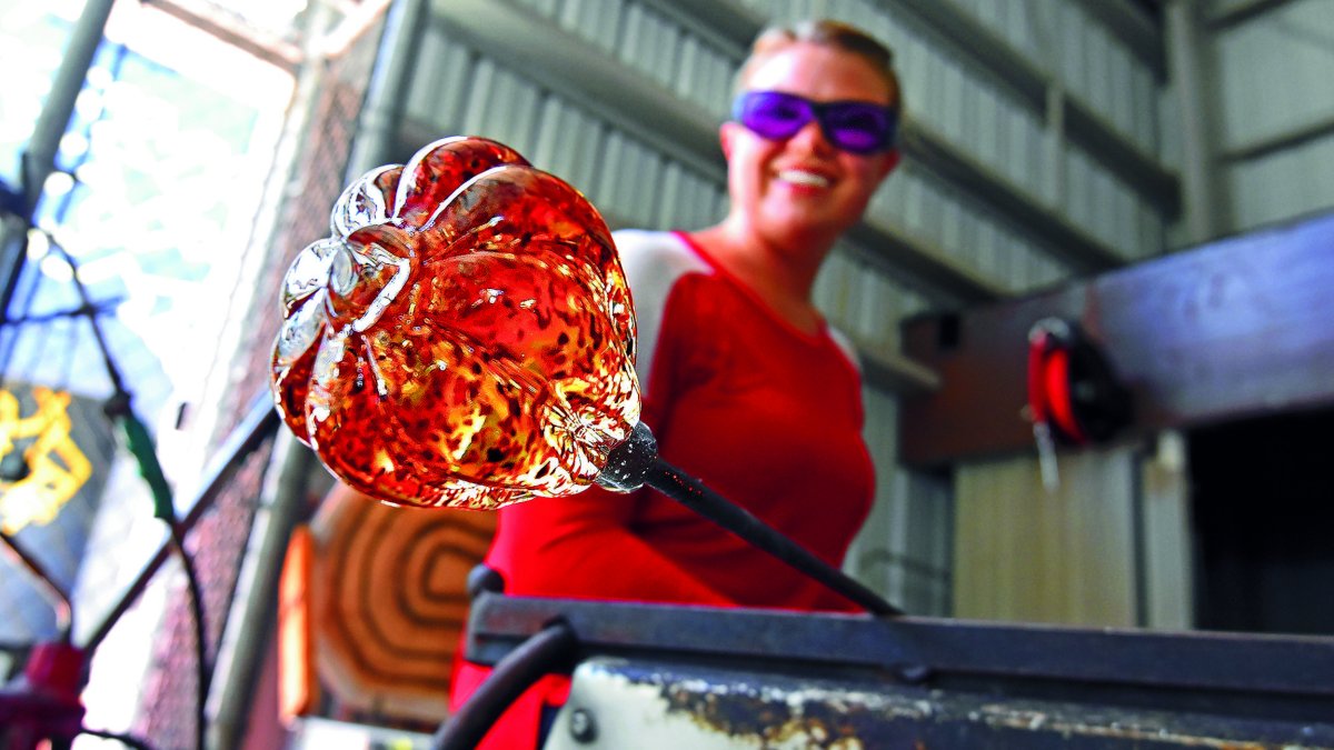 A woman holds an orange piece of art glass in a hot shop