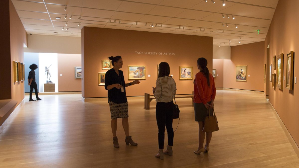 Three people talk in a gallery of paintings at the James Museum.