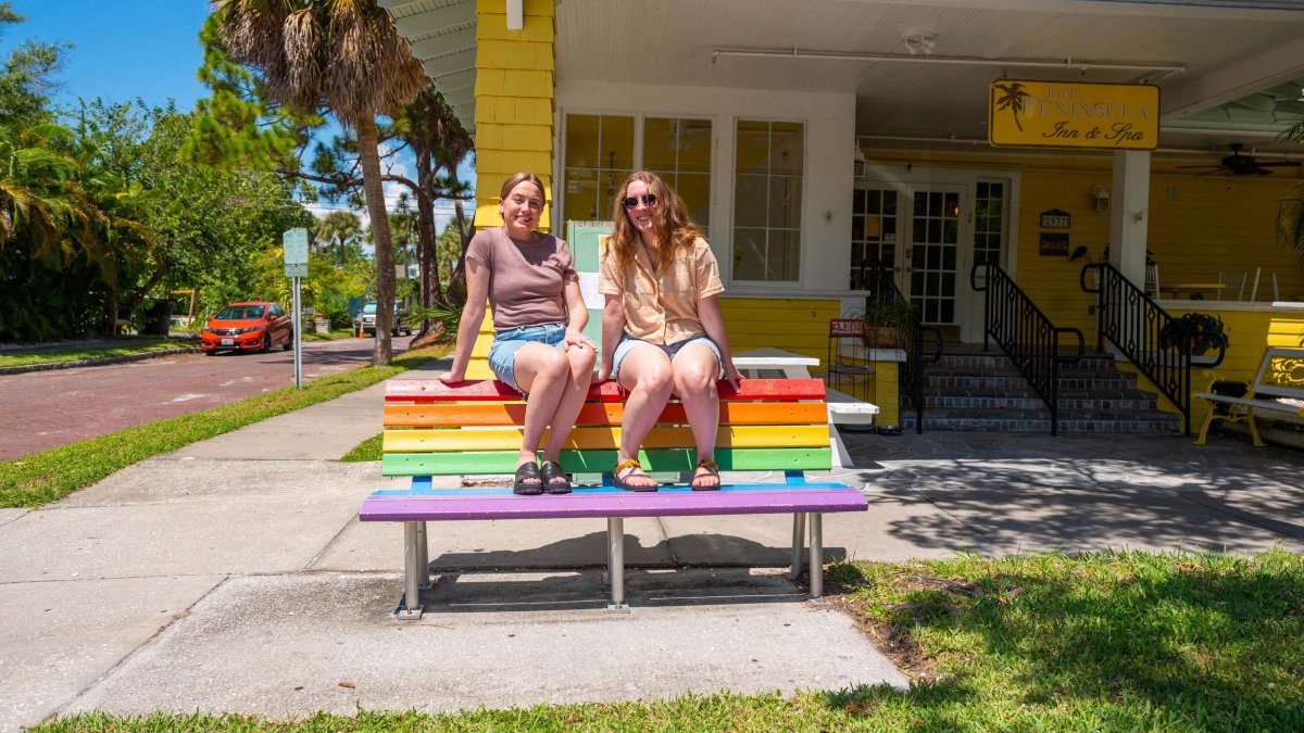 two women in shorts and tshirts sit on top of a bench painted in rainbow colors in front of a bright yellow inn next to a street