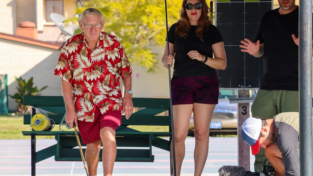 a man in a red hawaiian shirt and shorts plays shuffleboard with several other people