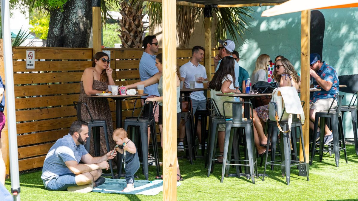 Adults and kids stand and sit at outdoor tables drinking beer and other beverages.