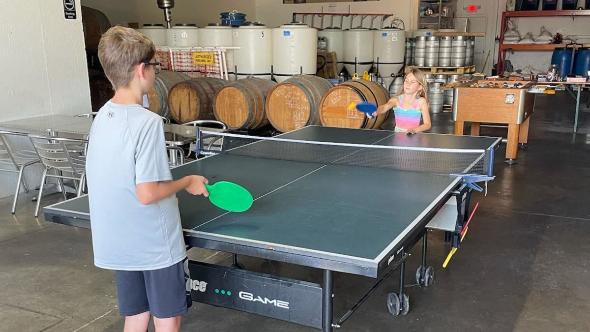 Two kids play table tennis with beer brewing equipment behind them