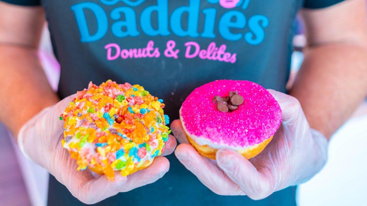 two donuts being held by a person wearing gloves, one donut has fruity pebbles the other has pink sugar crystals