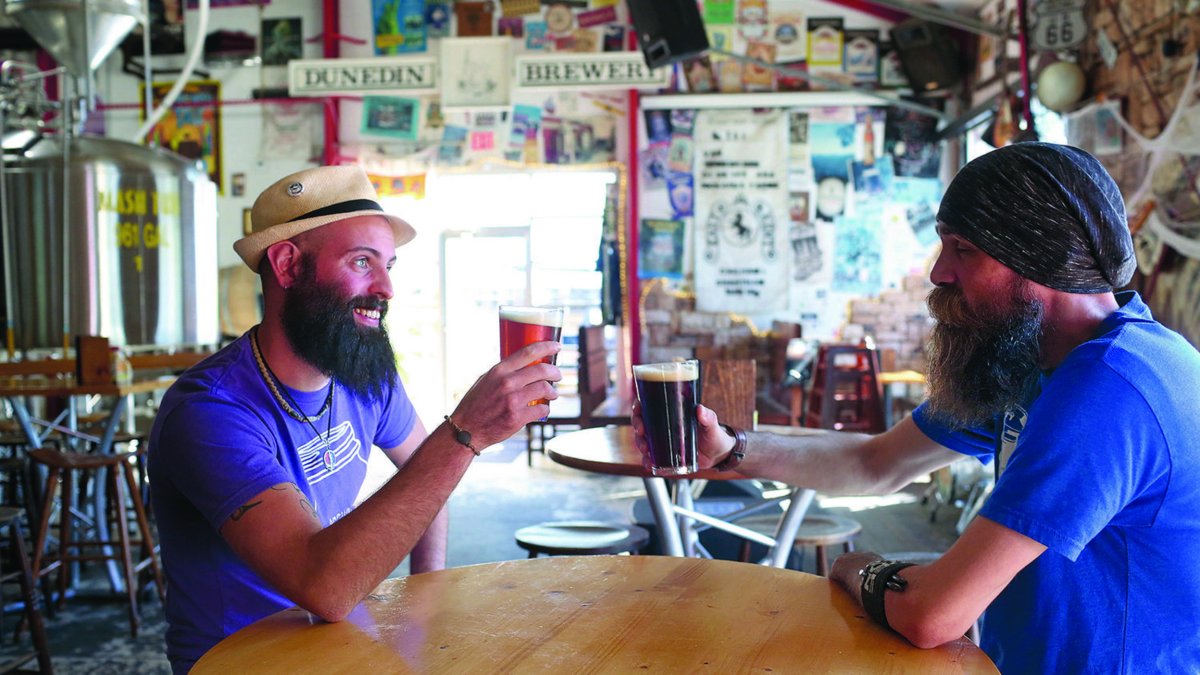 Two men in blue shirts toast with beer. Walls are covered with stickers.