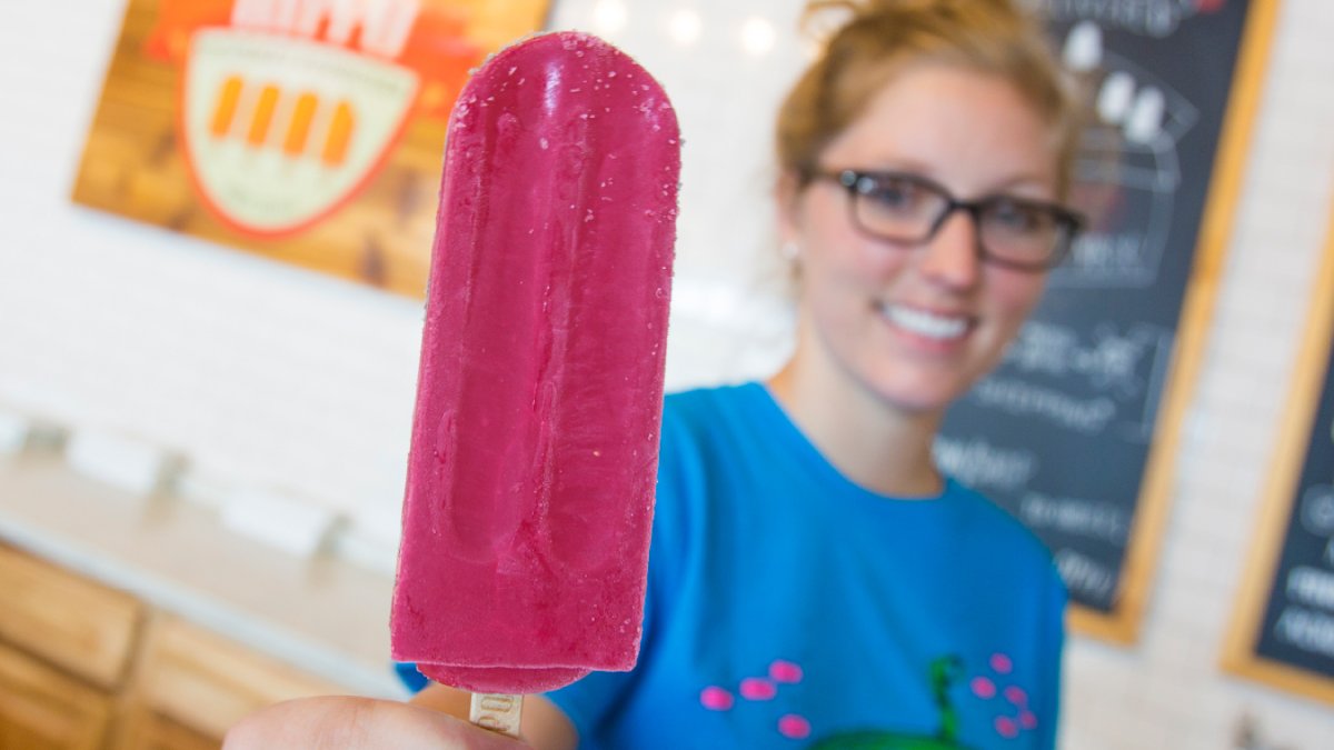 a hyppo popsicle employee in a blue shirt and glasses holds out a bright pink popsicle to a customer