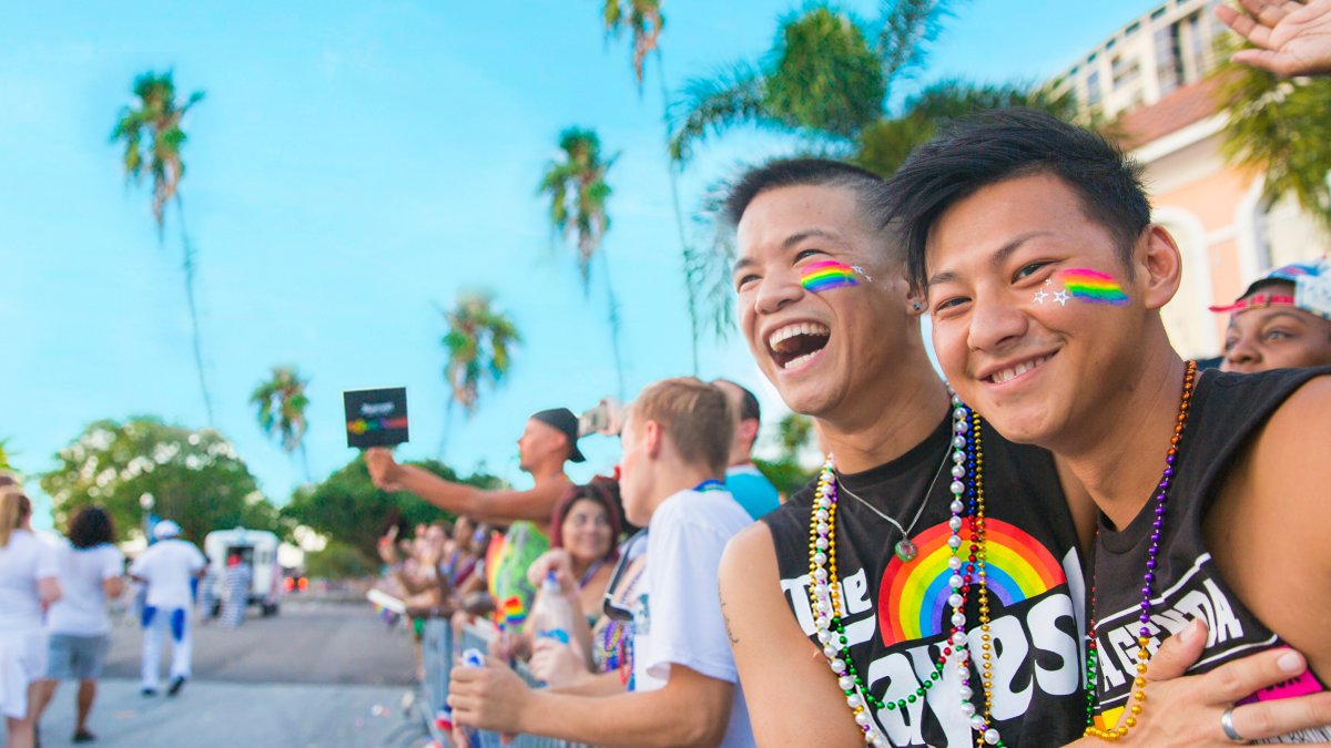 two young asian men in pride clothing smile as a parade goes by