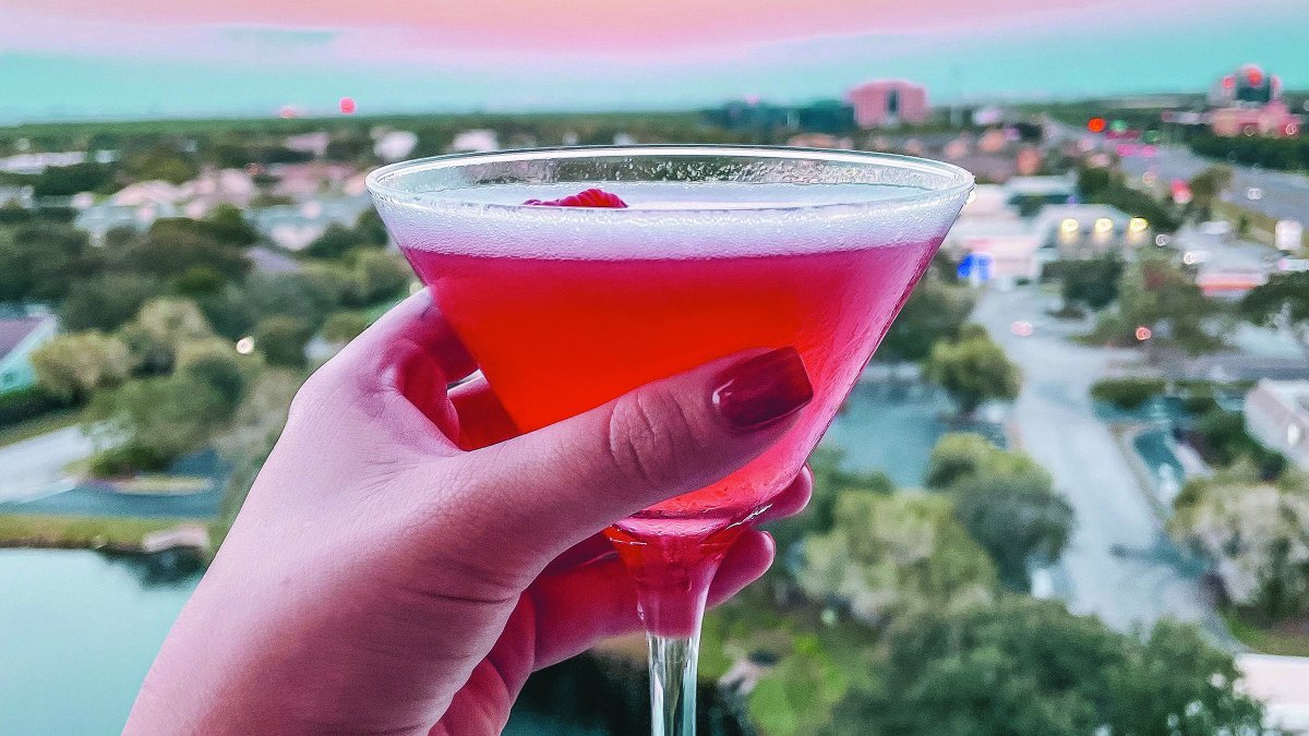 a manicured hand holding a pink cocktail with a cherry with a view of sunset and the Gulf from Vantage Rooftop Bar at the Karol Hotel