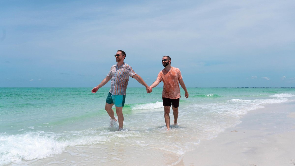 Two men hold hands while walking in shallow water on a pristine beach