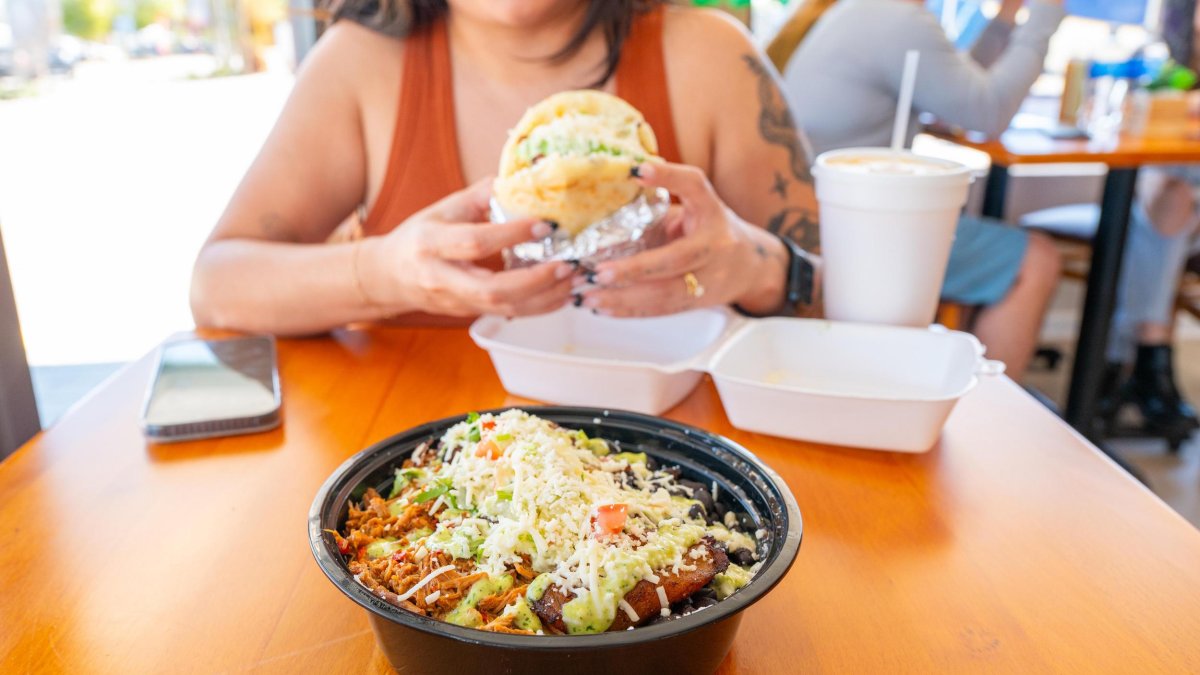 a woman in a tank top holds a Venezuelan arepa sandwich at a table, with a bowl of food in front of her
