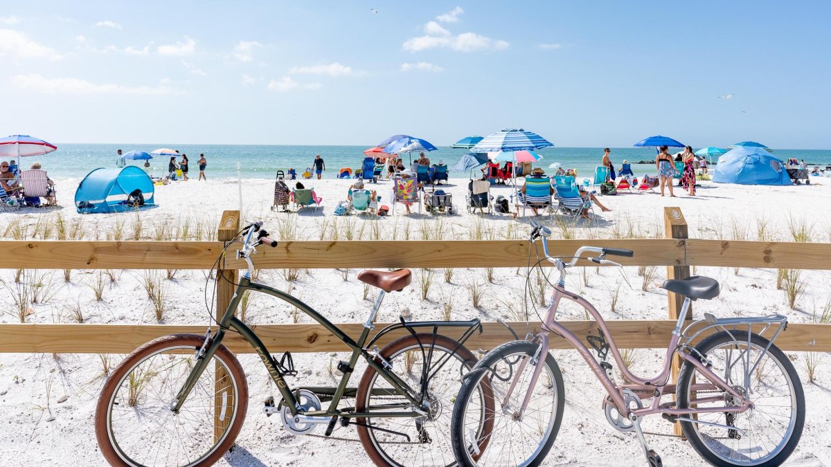 two bikes lean up against a wooden fence near a white-sand beach with umbrellas and people in chairs