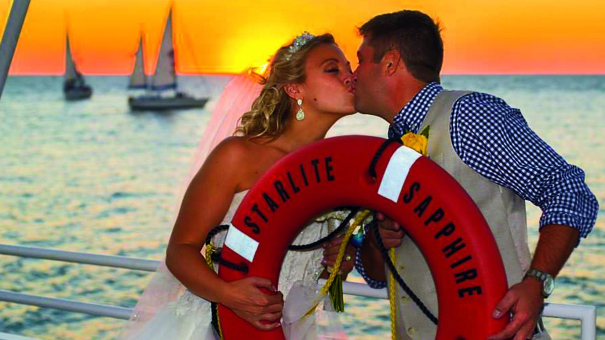 a bridal couple kiss while holding a large lifebuoy on StarLite Cruises with the sunset and sailboats behind them