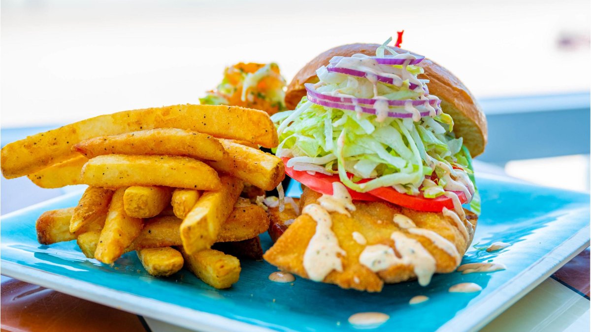 fried grouper sandwich with onions lettuce tomatoes and french fries on a bright blue plate