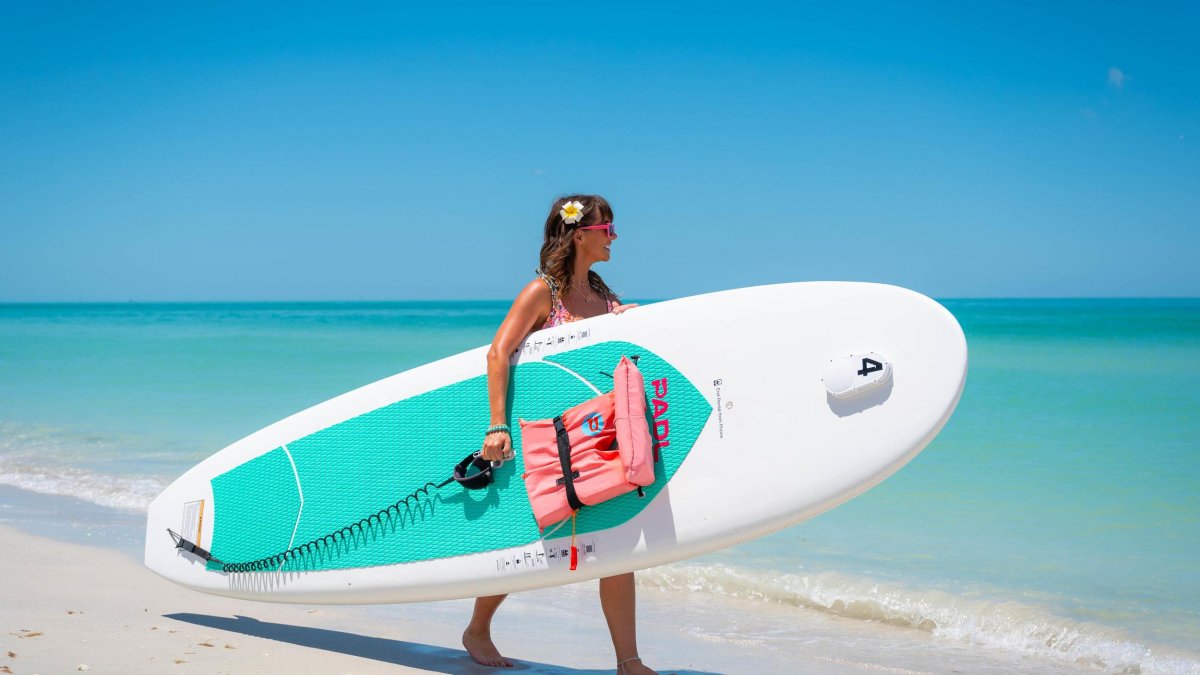 Woman walks along the ocean holding a stand-up paddleboard under her arm.