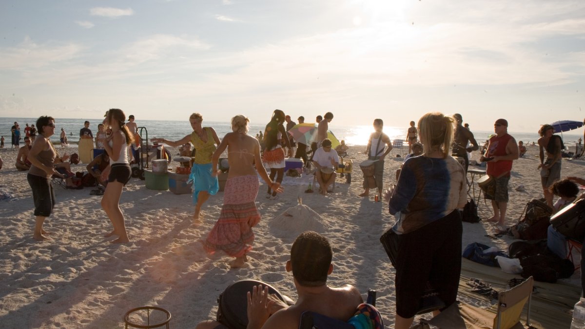 A group of beach-goers dance in the sand to drums on the beach at sunset.