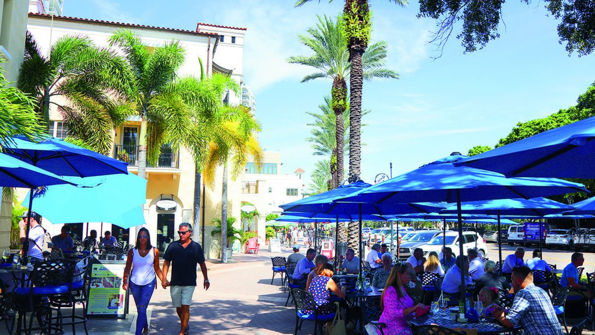 Outdoor restaurant with diners at tables under blue umbrellas and people walking by under palm trees and blue sky