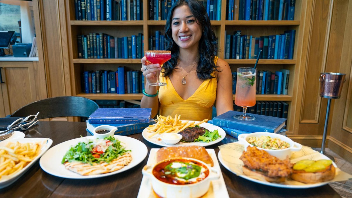 A woman in a yellow dress holds up a cocktail in front of plates of food with a bookshelf behind her