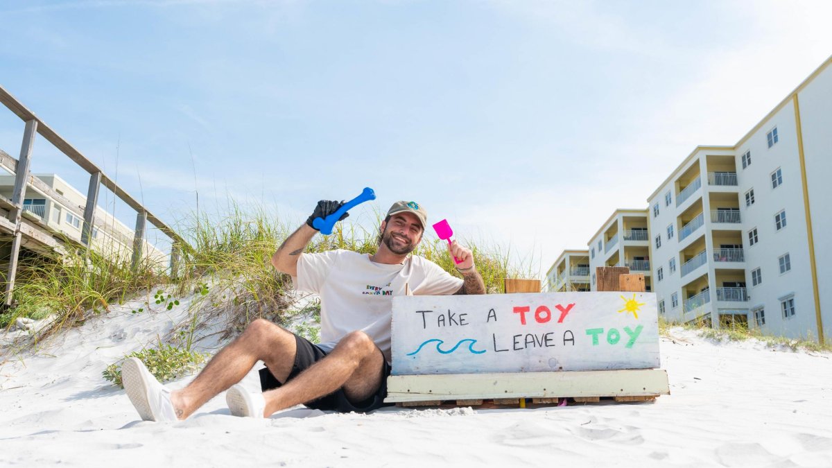 environmental influencer "Trash Caulin" sits near a dune with some plastic beach toys and a sign that says "take a toy, leave a toy"
