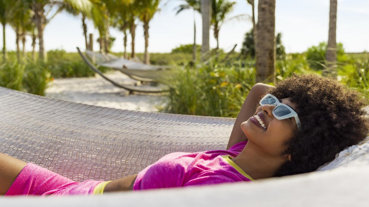 Woman with sunglasses laying in a hammock with the beach and palm trees in the background