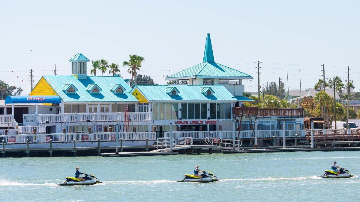 Exterior of a waterfront restaurant and dock with jet skiers going by.