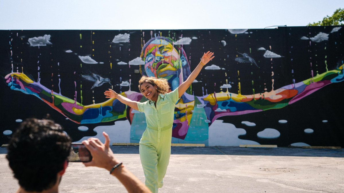 a person taking a photo of a woman wearing a lime green jumpsuit with her arms outstretched in front of a mural of a black woman with her arms outstretched