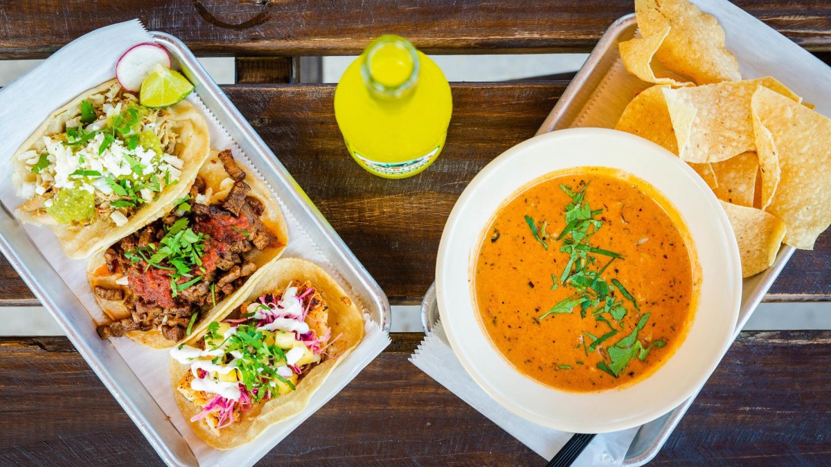 a tray with three kinds of tacos next to a tray with mexican soup and chips, with a yellow soda in the middle