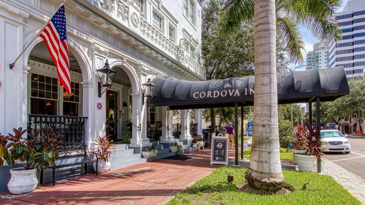 the white facade of the Cordova Inn with a huge black awning and an American flag, next to large palm trees, brick walkway and grass
