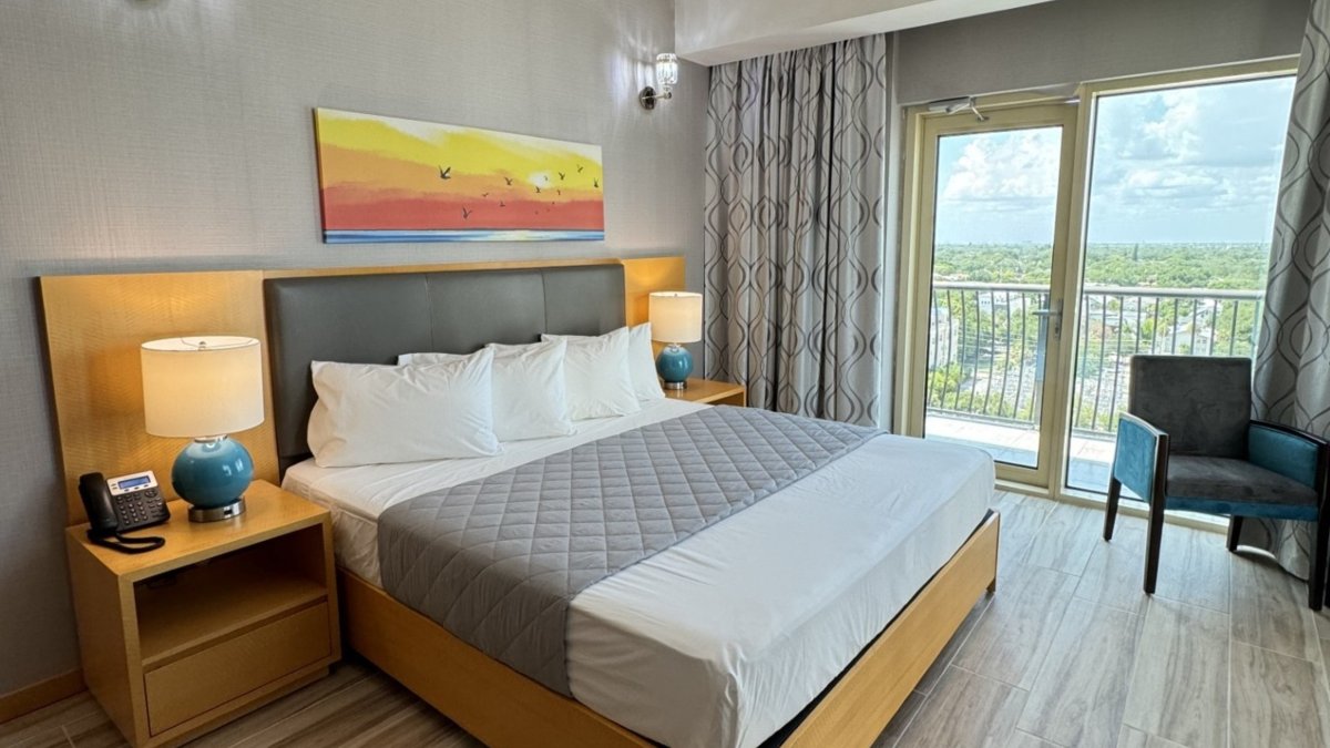 image of hotel room with clean modern furniture and white bedding with a glass sliding door with a view of downtown St. Pete