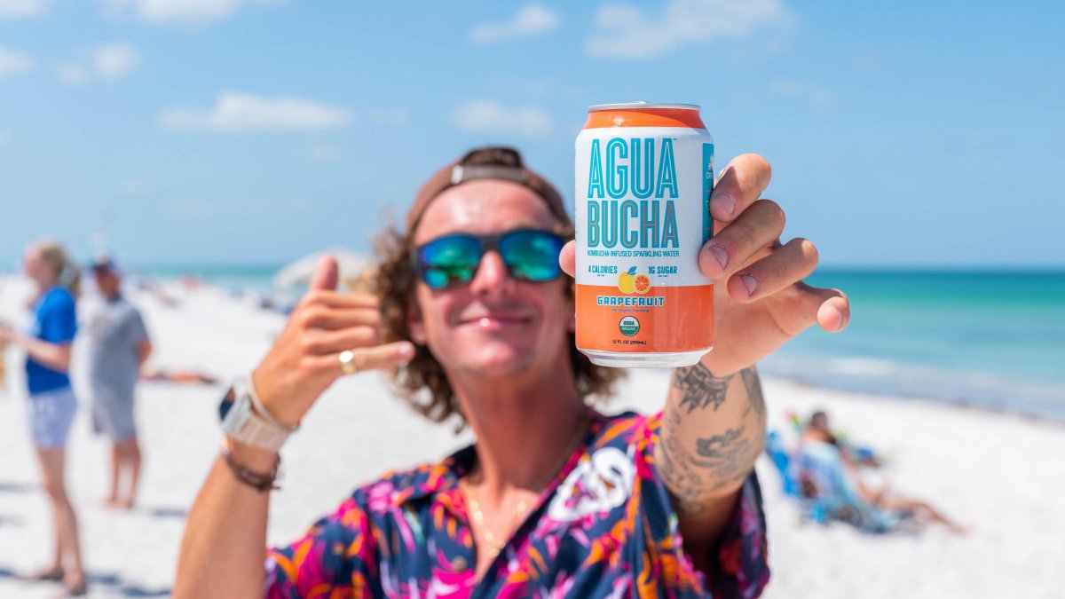 a person on the beach with a baseball cap on backwards, mirrored sunglasses and a tropical shirt holds out a can of kombucha