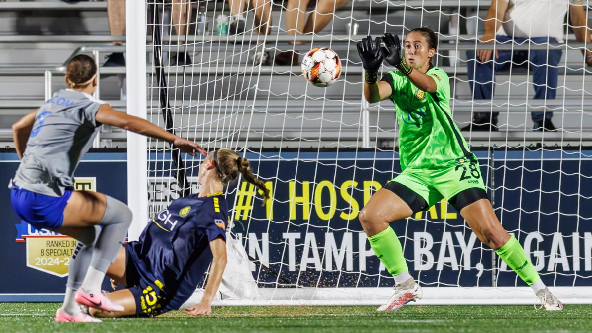 a goalie in a bright green shirt blocks the ball at a women's soccer game, with two players nearby and people in the stands; Photo credit: Mike Carlson