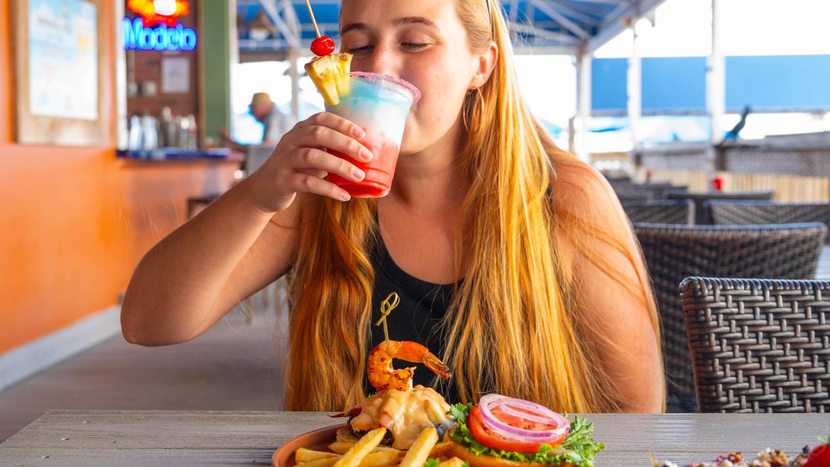 a woman sipping a red white and blue frosty cocktail at an outdoor restaurant with a sandwich and fries in front of her