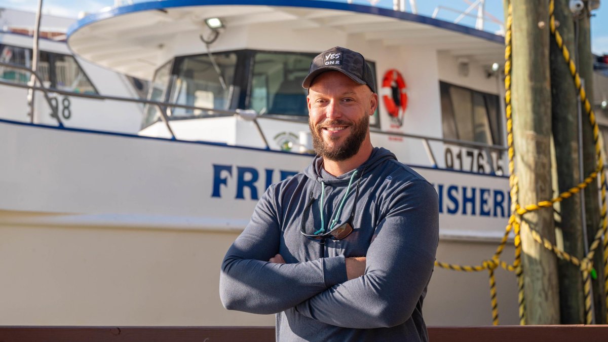 a smiling man with a beard wearing a baseball cap standing in front of a large fishing boat