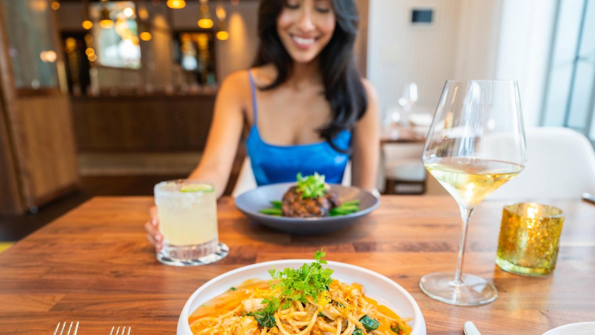 a smiling woman in a bright blue dress sits across from a bowl of pasta at a restaurant