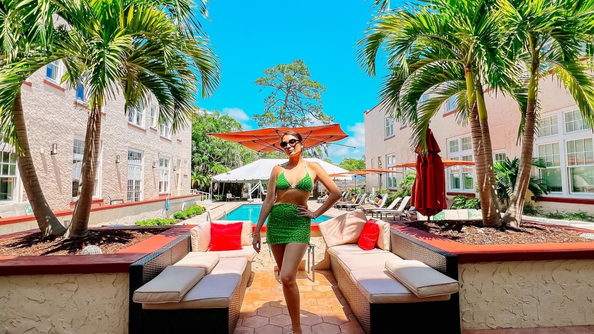 a woman in a bright green bathing suit posing in front of benches and a pool near palm trees at the Fenway Hotel