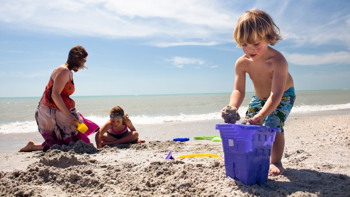 Ein kleiner Junge schüttet Sand in einen großen lila Eimer in Form einer Sandburg, während seine Mutter und seine Schwester hinter ihm am Strand im Sand spielen