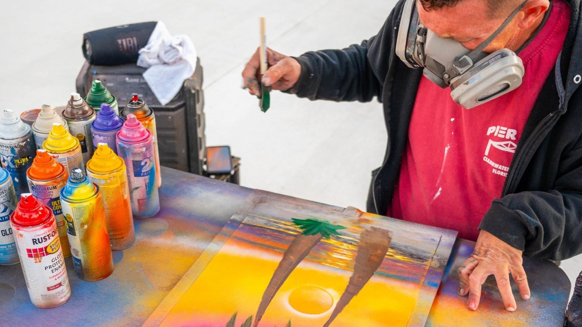 an artist wearing a heavy-duty mask uses airbrushed paints to create a palm tree painting at the Sunsets at Pier 60 celebration