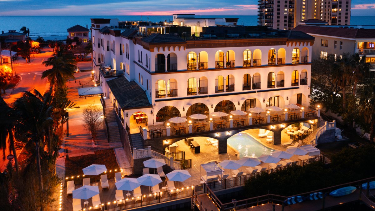 an aerial exterior view of the Hotel Zamora lit up at night, with the beach visible behind the hotel, and tables with umbrellas in the forefront along the bayside of the building