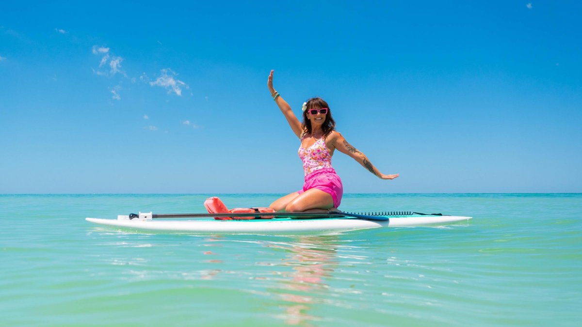 a woman dressed in pink clothing and sunglasses smiles as she sits on top of a paddleboard at the beach