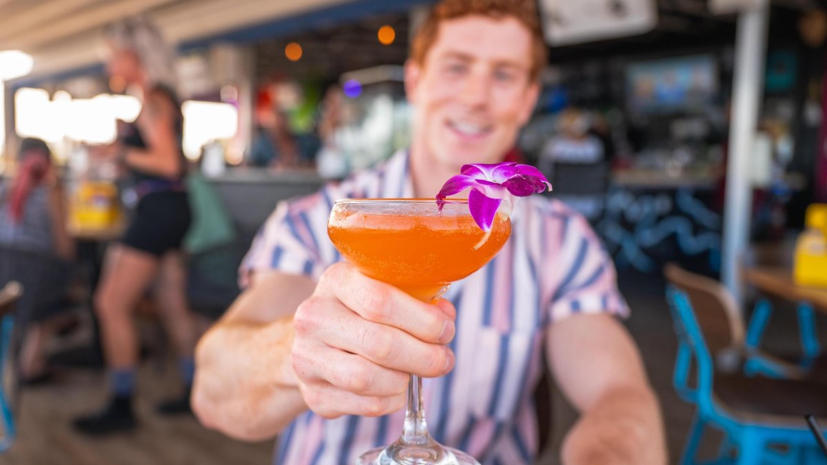 man holding colorful cocktail up with flower on rim at an outdoor restaurant
