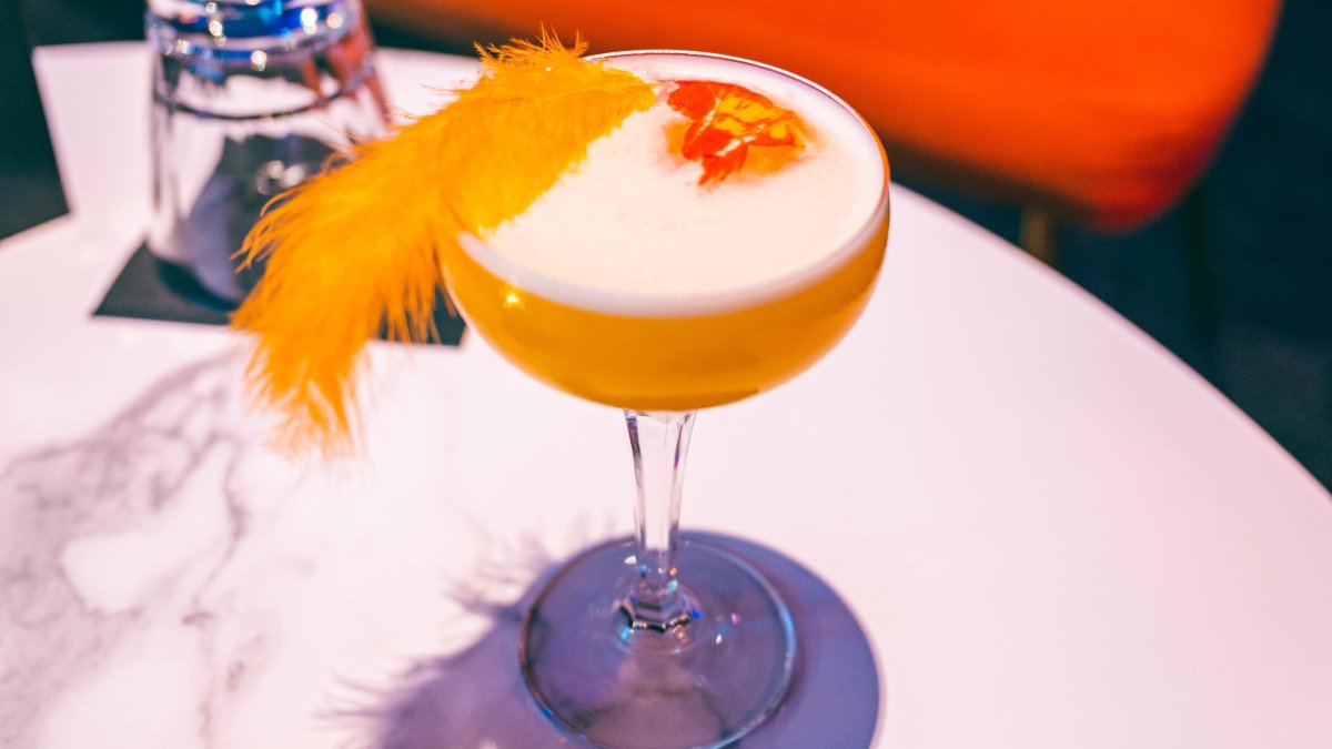 An orange-colored craft cocktail with a feather garnish sits on a table