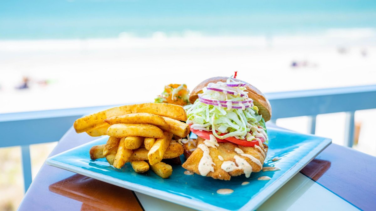 a fish sandwich and fries on a table that looks like a surfboard at Caddy's Madeira Beach, with a view of the beach in the distance