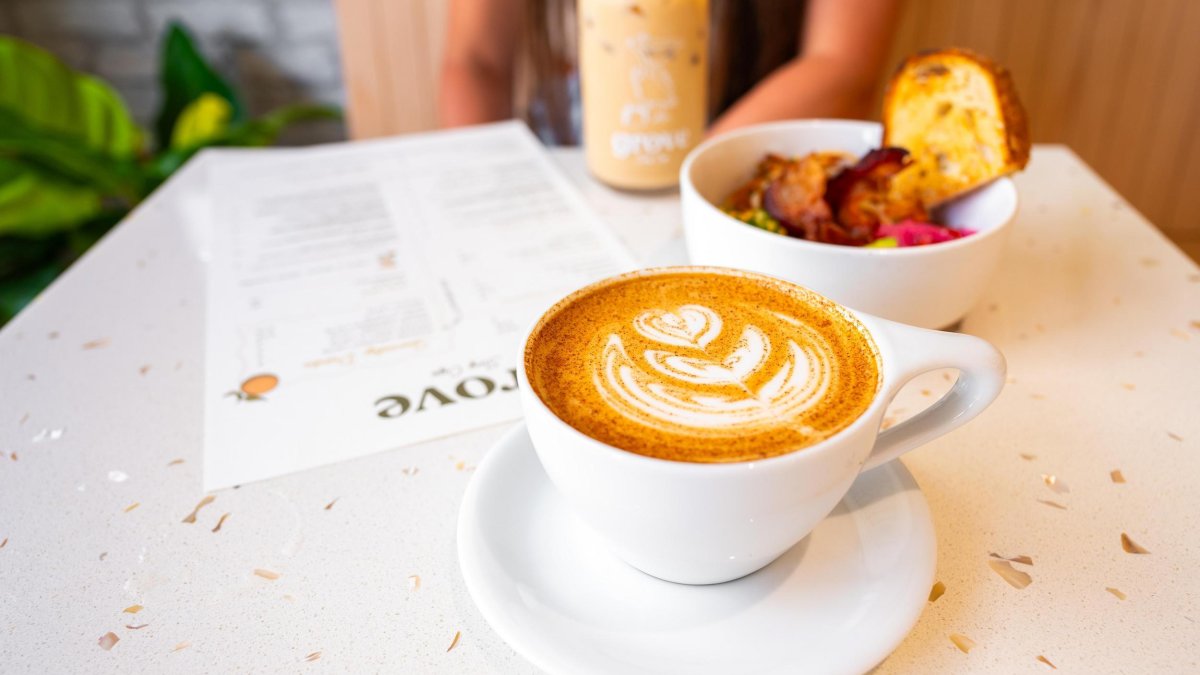 a hot latte with a pretty design next to an iced coffee and a breakfast bowl on a table at Grove Surf Cafe